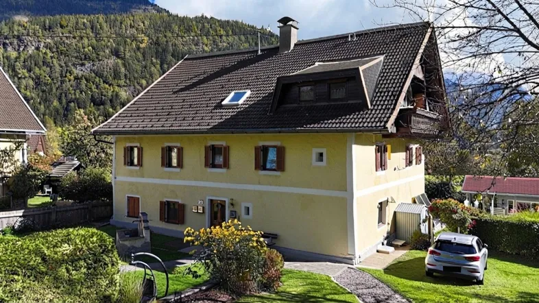 Traditionelles Haus mit gelber Fassade, Satteldach und Bergblick in ländlicher Umgebung.