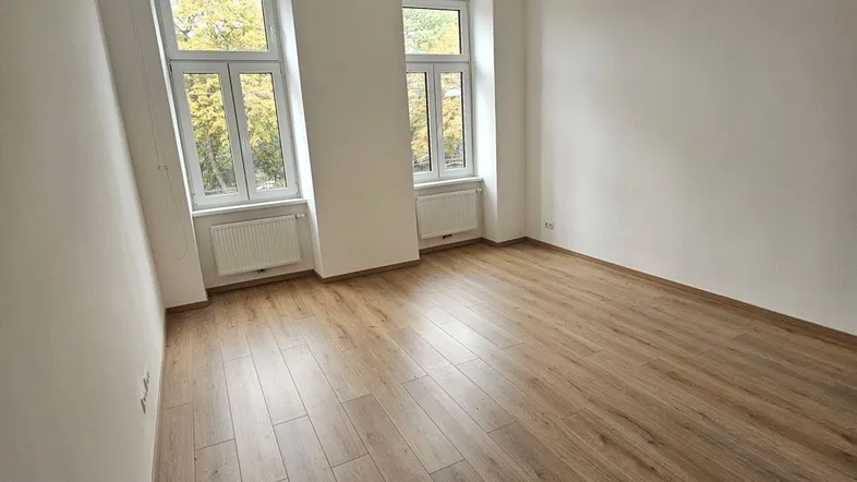 Helles, leeres Zimmer mit Laminatboden und zwei hohen Fenstern, die viel Tageslicht hereinlassen.