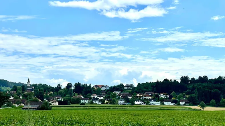 Panoramablick auf ein malerisches Dorf mit Kirche, umgeben von grünen Feldern und bewaldeten Hügeln unter blauem Himmel.