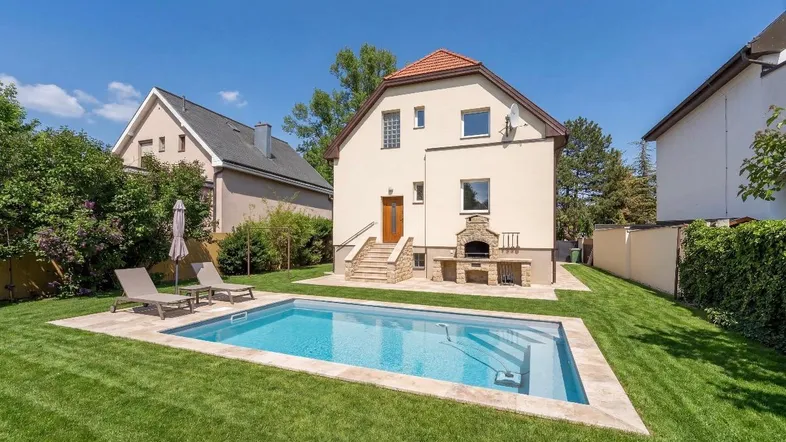 Haus mit gepflegtem Garten, Pool und Grillbereich unter blauem Himmel.