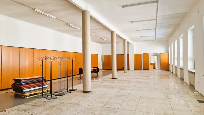 Großer, heller offener Raum mit hohen Decken, Säulen, Fliesenboden und integrierten Holzschränken entlang der Wand.