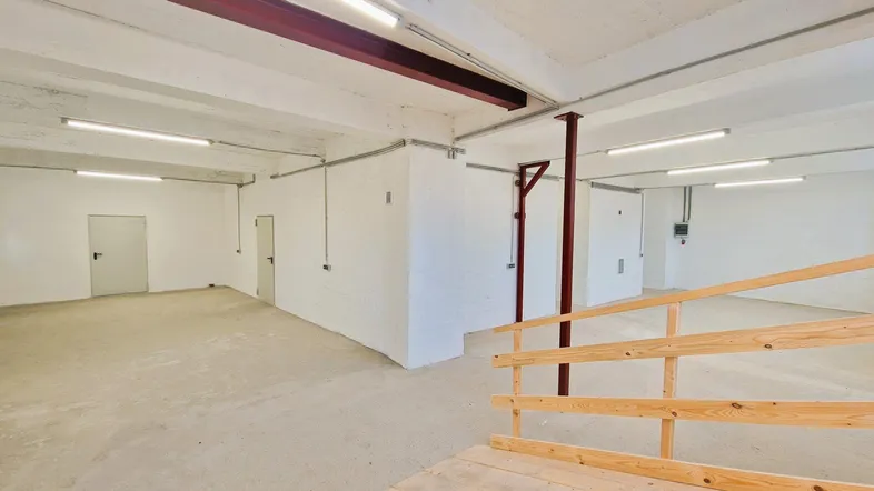 Geräumiger, leerer Lagerraum mit Betonboden, weißen Wänden, sichtbaren Stahlträgern und einer Holzrampe für den Zugang.