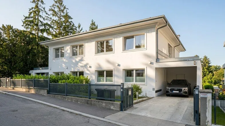 Modernes weißes Einfamilienhaus mit Carport und gepflegter Außenanlage in grüner Umgebung.
