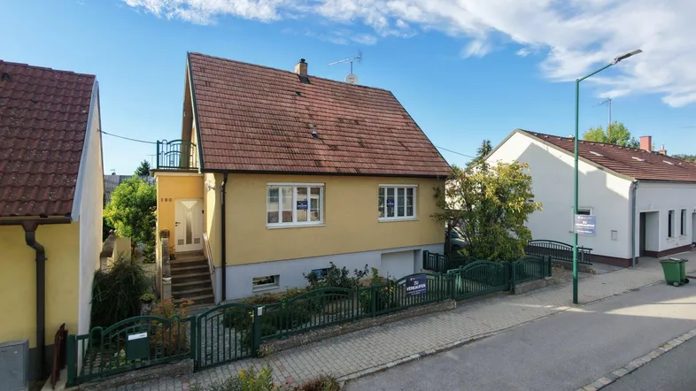 Gelbes Einfamilienhaus mit rotem Ziegeldach und Vorgarten an einer ruhigen Straße.