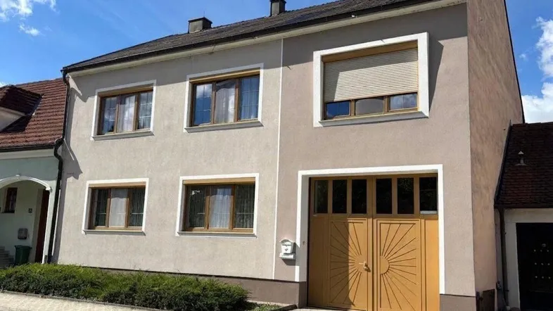 Helles Einfamilienhaus mit beiger Fassade, braunen Fenstern und Rollläden.