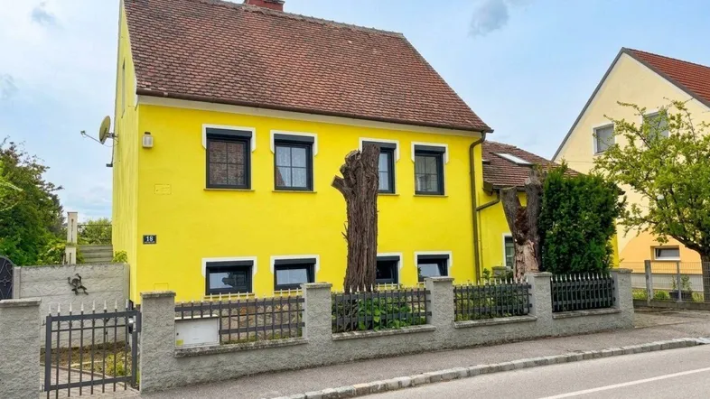 Gelbes Einfamilienhaus mit rotem Ziegeldach und Vorgarten an einer ruhigen Straße.