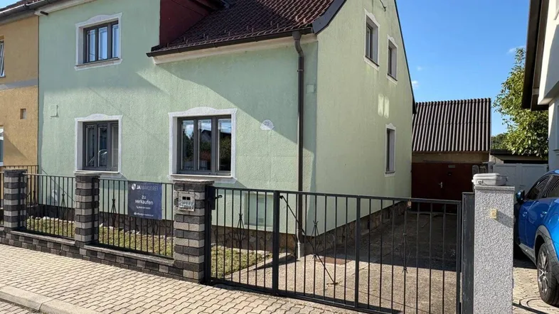 Hausfassade in hellem Grün mit Zaun und Einfahrt unter blauem Himmel.