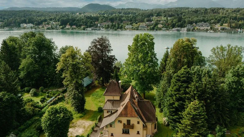 Luftaufnahme eines historischen Gebäudes mit weitläufigem Garten und Blick auf den See und die Berge.
