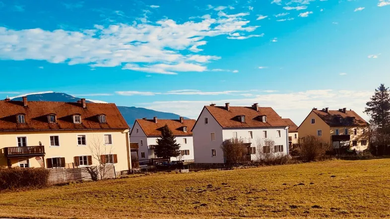 Mehrere Wohnhäuser in einer ländlichen Umgebung mit Bergen im Hintergrund unter blauem Himmel.