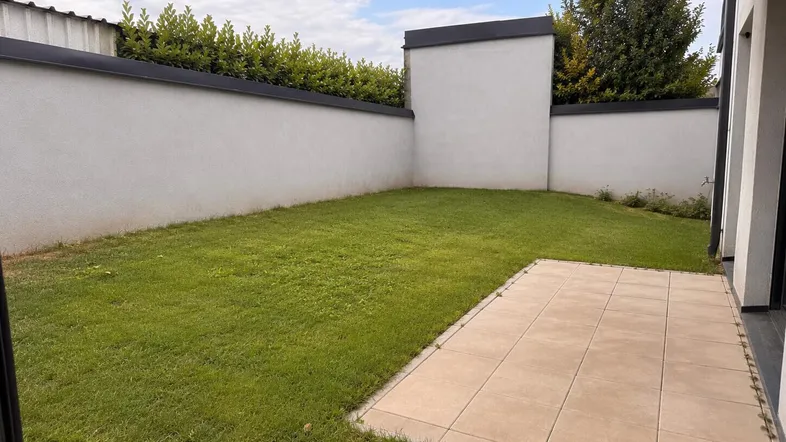 Kleiner, gepflegter Gartenbereich mit Rasenfläche und gepflasterter Terrasse, umgeben von einer Hecke.