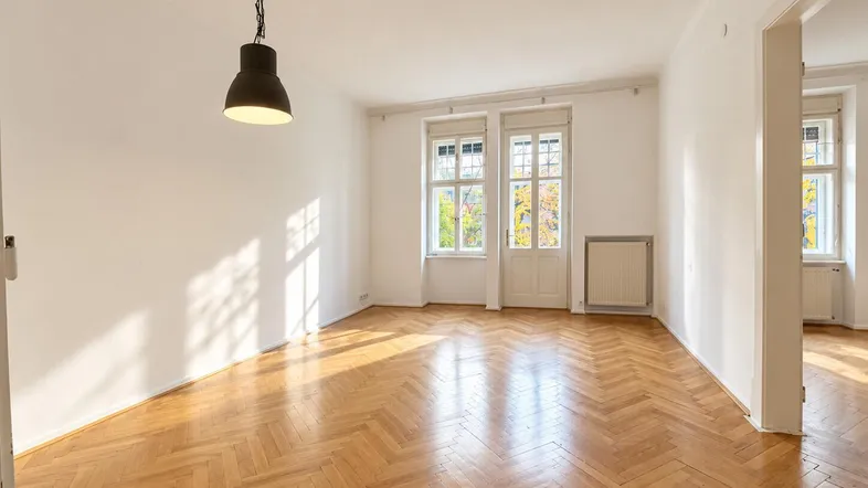 Helles, leeres Zimmer mit Fischgrätparkett und großen Fenstern, das viel Tageslicht hereinlässt.
