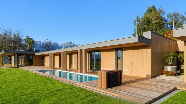 Moderne Villa mit Holzfassade, langem Pool und weitläufigem Garten vor alpiner Bergkulisse.