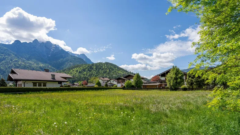 Weitläufige grüne Landschaft mit Häusern und majestätischen Bergen im Hintergrund unter blauem Himmel.