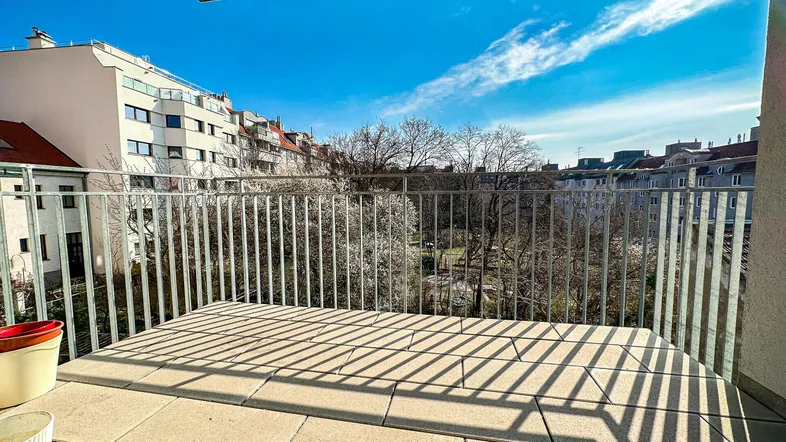 Sonniger Balkon mit Fliesenboden und Blick auf grüne Bäume sowie umliegende Gebäude.