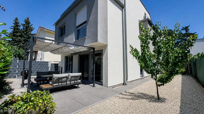 Moderne Hausfassade mit großer gepflasterter Terrasse, Pergola und Kiesgarten.