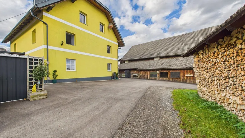 Gelbes Wohnhaus mit weißem Streifen und angrenzendem Holzschuppen auf einem ländlichen Grundstück.