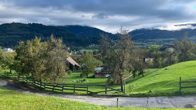 Panoramablick über eine grüne Hügellandschaft mit vereinzelten Häusern und Bergen im Hintergrund unter bewölktem Himmel.