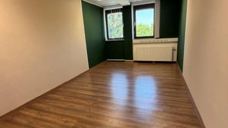 Leeres Büro mit Laminatboden, abgehängter Decke und zwei Fenstern, die einen Blick ins Grüne bieten.