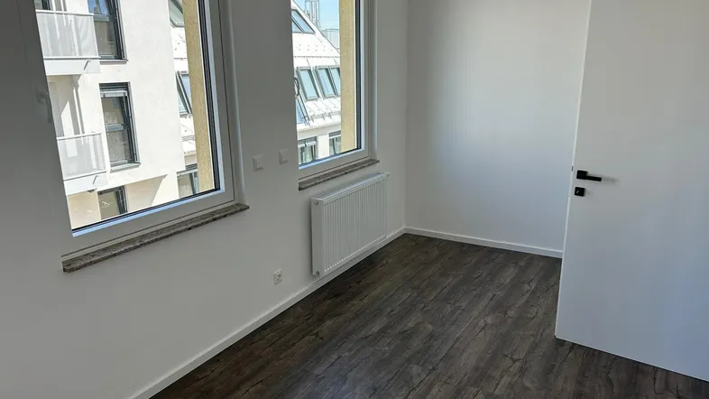 Helles Zimmer mit dunklem Laminatboden und Blick auf das angrenzende Gebäude.