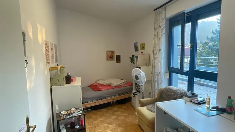 Helles Schlafzimmer mit Parkettboden, Schreibtisch und Zugang zu einem Balkon.