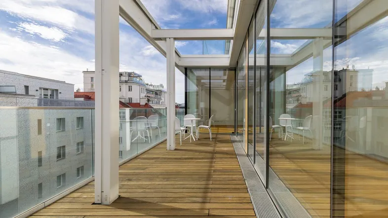 Moderne Dachterrasse mit Holzboden, Glasgeländer und Sitzgelegenheiten unter blauem Himmel.