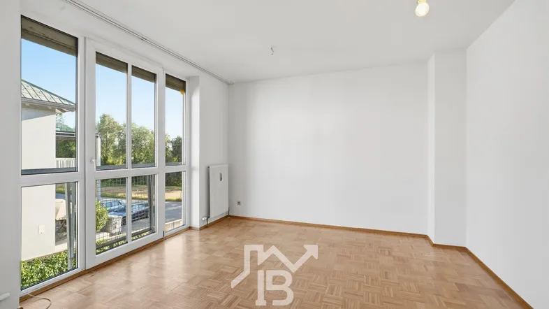 Helles, leeres Wohnzimmer mit Parkettboden und großen Fenstern, die viel Tageslicht hereinlassen.