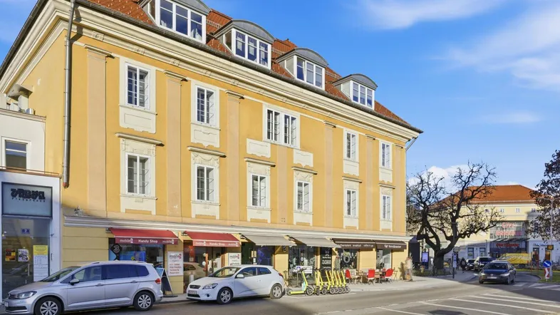 Historisches Gebäude mit gelber Fassade, roten Dachziegeln und Geschäften im Erdgeschoss an einer belebten Straße.