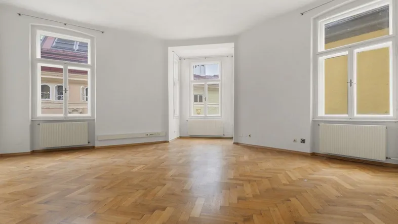 Geräumiger Wohnraum mit Fischgrätparkett, hohen Fenstern und viel Tageslicht mit Blick auf die Stadt.