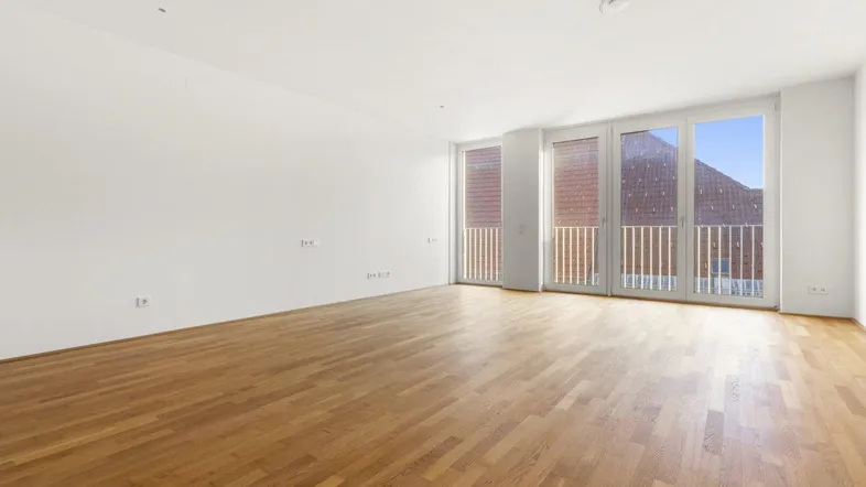 Heller, leerer Raum mit großen Fenstern, Holzboden und Zugang zu einem französischen Balkon.