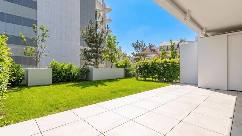 Grüne Gartenfläche mit gepflasterter Terrasse und Blick auf umliegende Gebäude.