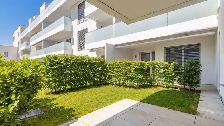 Gepflegter Gartenbereich mit Rasenfläche und Hecke, angrenzend an eine moderne Wohnanlage.