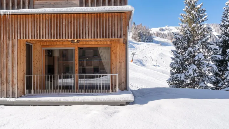 Modernes Holzgebäude mit Balkon und direktem Blick auf die verschneite Skipiste und die Berge.