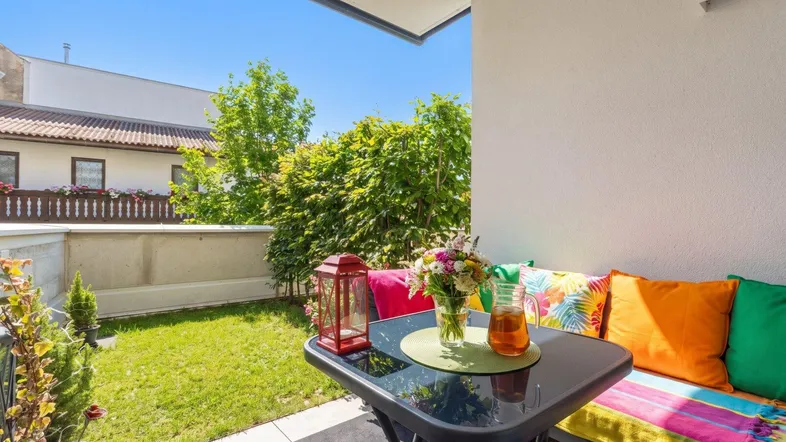 Sonnige Terrasse mit bunten Sitzgelegenheiten und kleinem Gartenbereich, umgeben von grünen Hecken.