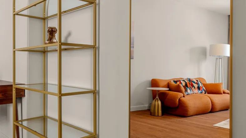Spiegelung eines hellen Wohnzimmers mit modernem Sofa und Stehlampe, ergänzt durch ein goldenes Regal.
