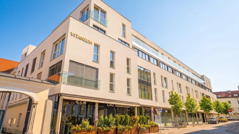 Modernes Gebäude mit weißer Fassade, großen Fenstern und Geschäften im Erdgeschoss an einer belebten Straße.