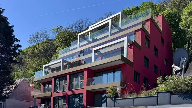 Modernes Gebäude mit roter Fassade, gestuften Terrassen und Glasgeländern, umgeben von viel Grün.