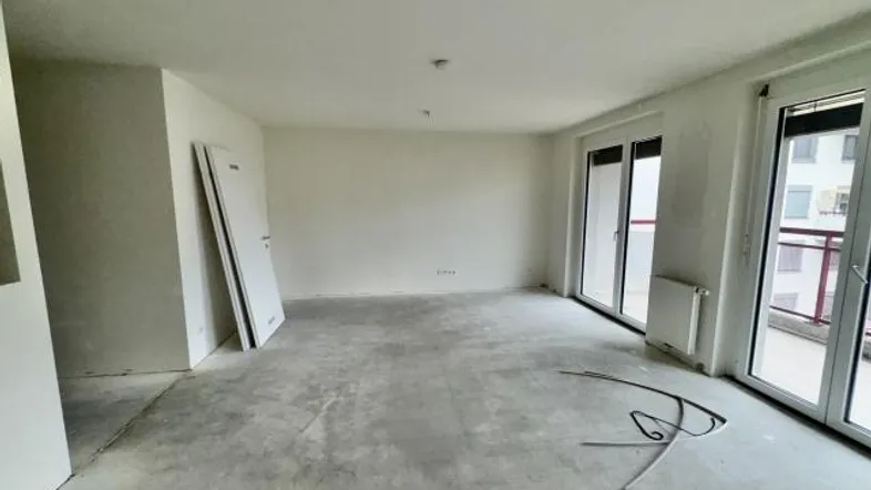 Heller, unfertiger Raum mit großen Fenstern und Zugang zu einem Balkon, bereit für den Ausbau.