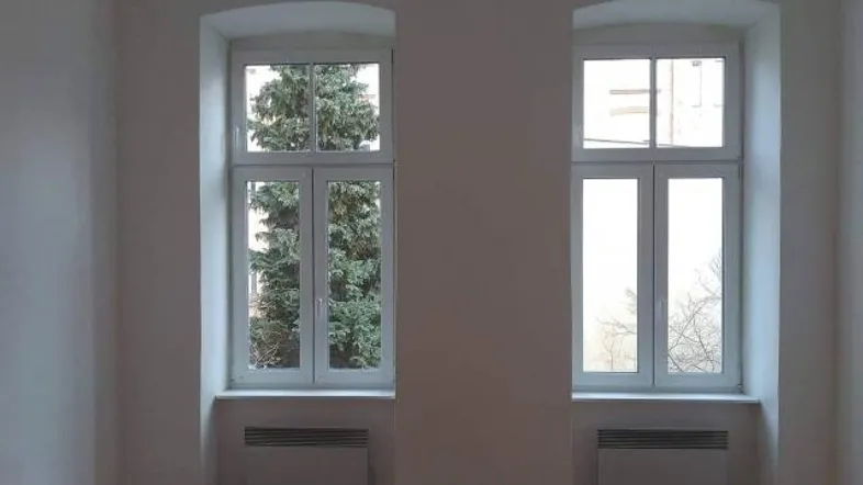 Heller Raum mit zwei hohen Fenstern, Holzboden und Blick ins Grüne.