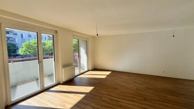 Helles, leeres Wohnzimmer mit Laminatboden und Zugang zu einem Balkon, der viel Tageslicht hereinlässt.