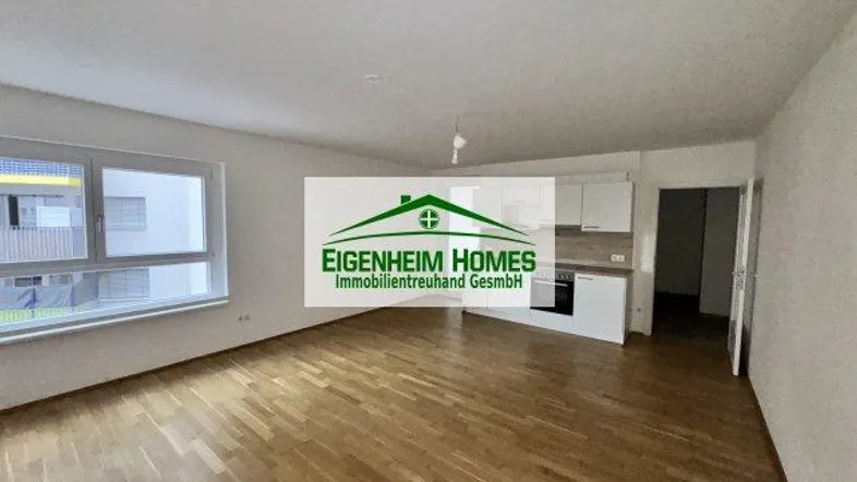 Heller Wohnbereich mit Parkettboden, großen Fenstern und integrierter Küchenzeile in einem Neubau.