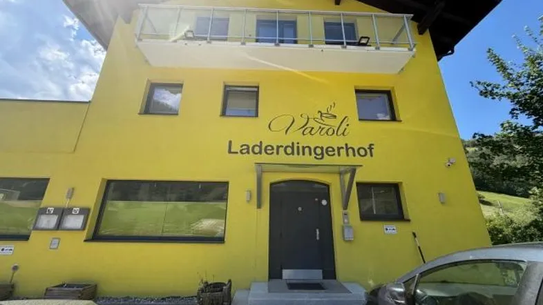 Gelbes Gebäude mit Balkon und Eingangsbereich, umgeben von grüner Landschaft unter blauem Himmel.