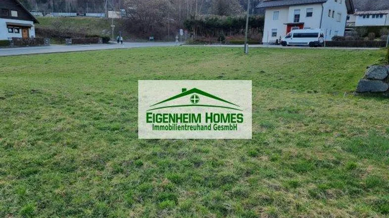 Blick auf ein grünes Grundstück mit Häusern im Hintergrund und bewaldeten Hügeln unter bewölktem Himmel.