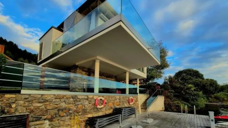 Modernes Gebäude mit Glasbalkonen und Holzterrasse direkt am Seeufer unter blauem Himmel.