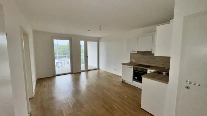 Helles Wohnzimmer mit integrierter Küchenzeile, Holzboden und Zugang zu einem Balkon.