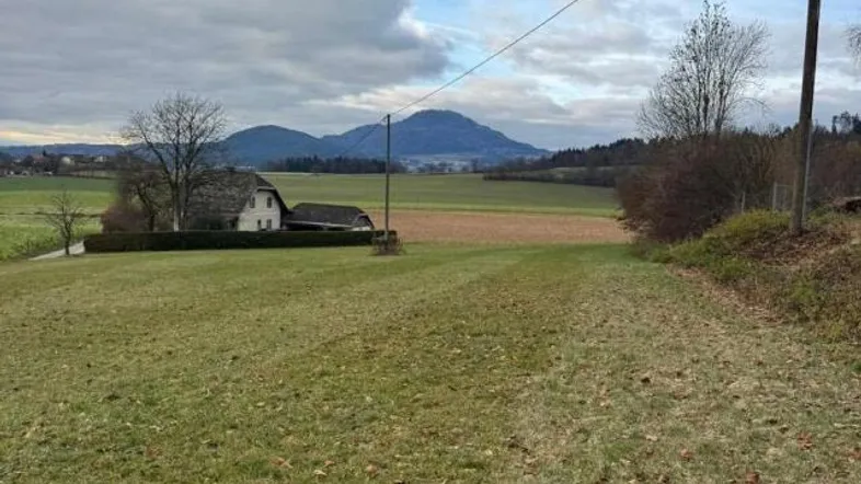 Weitläufige grüne Landschaft mit einem Haus im Hintergrund und Bergen am Horizont unter bewölktem Himmel.