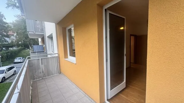 Heller Balkon mit Fliesenboden und Blick auf die Straße sowie umliegende Bäume.