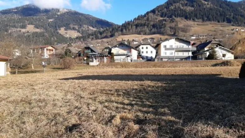Blick über ein trockenes Feld auf eine Siedlung mit Häusern und bewaldeten Bergen im Hintergrund.