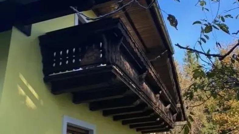 Traditioneller Holzbalkon an einem Haus mit gelber Fassade unter blauem Himmel und Bäumen.