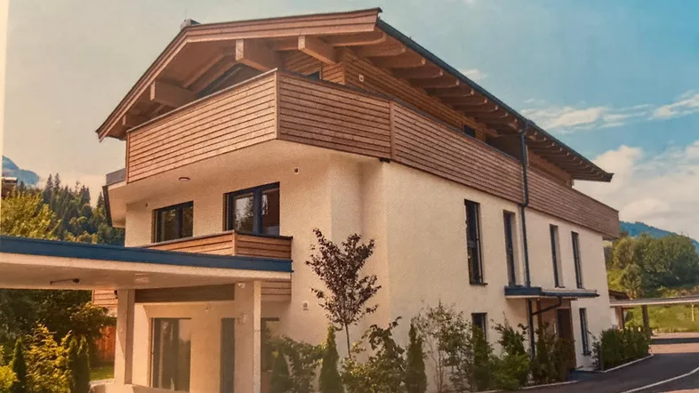 Modernes Wohnhaus mit Holzverkleidung, großem Balkon und Carport in alpiner Umgebung.