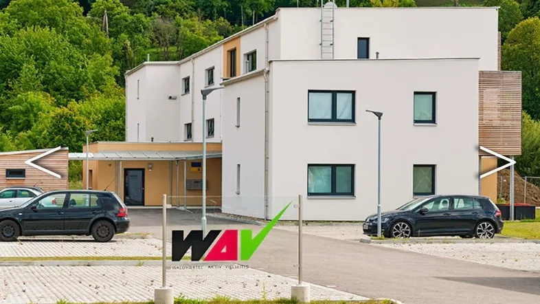 Modernes Mehrfamilienhaus mit weißer und beiger Fassade, umgeben von grüner Landschaft und Parkplätzen.